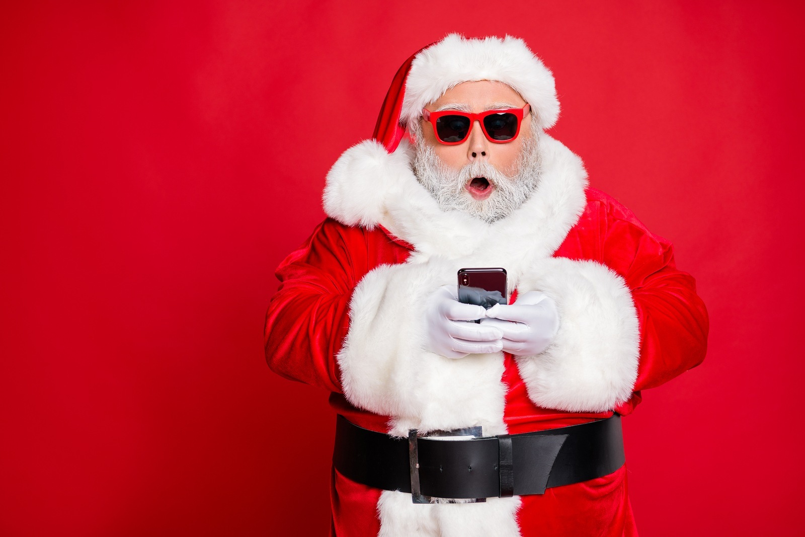 Portrait of impressed Christmas father in hat using cell phone device screaming omg wearing read about festive events eyeglasses eyewear belt isolated over red background