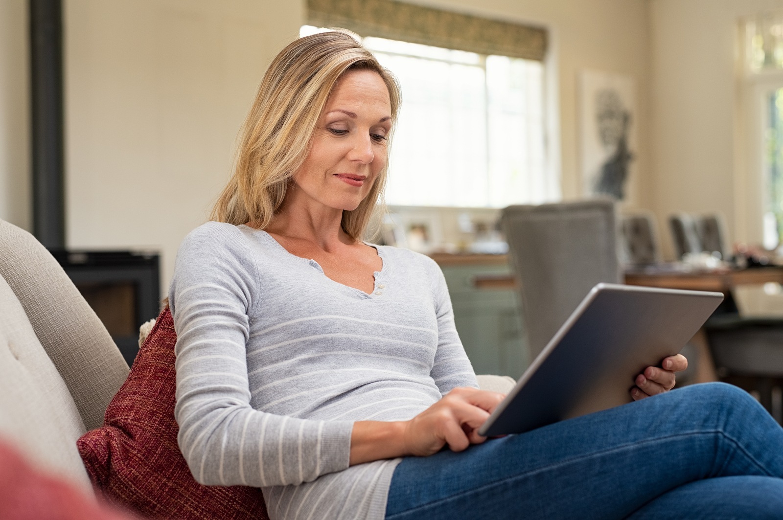 Beautiful mature woman sitting on couch and using digital tablet. Smiling lady browsing internet over tablet at home. Middle age woman holding computer at home.