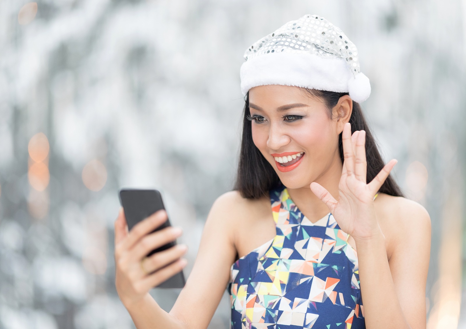 Beautiful toothy smiling young woman in white Santa's hat using smartphone doing facetime with family against blur background.Christmas concept.