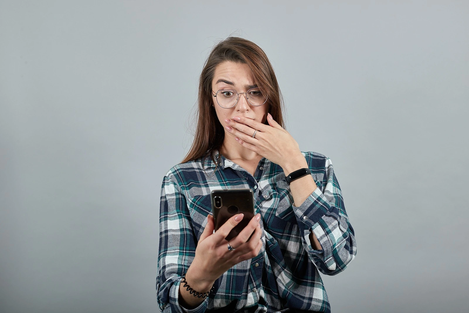 Young brunette girl blue green in checked shirt on grey background, shocked woman with glasses is talking on phone that looks like an iPhone