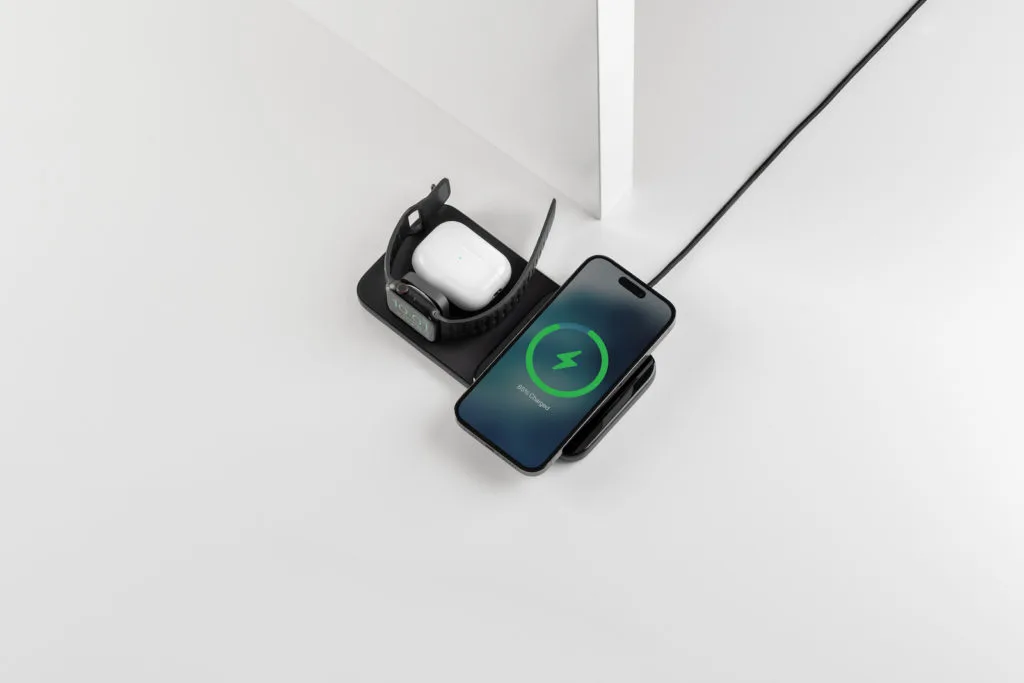 Das Nomad Base One Max 3-in-1 Ladegerät in Silber auf einem Tisch, während es gleichzeitig ein iPhone über MagSafe, eine Apple Watch und AirPods kabellos auflädt.
