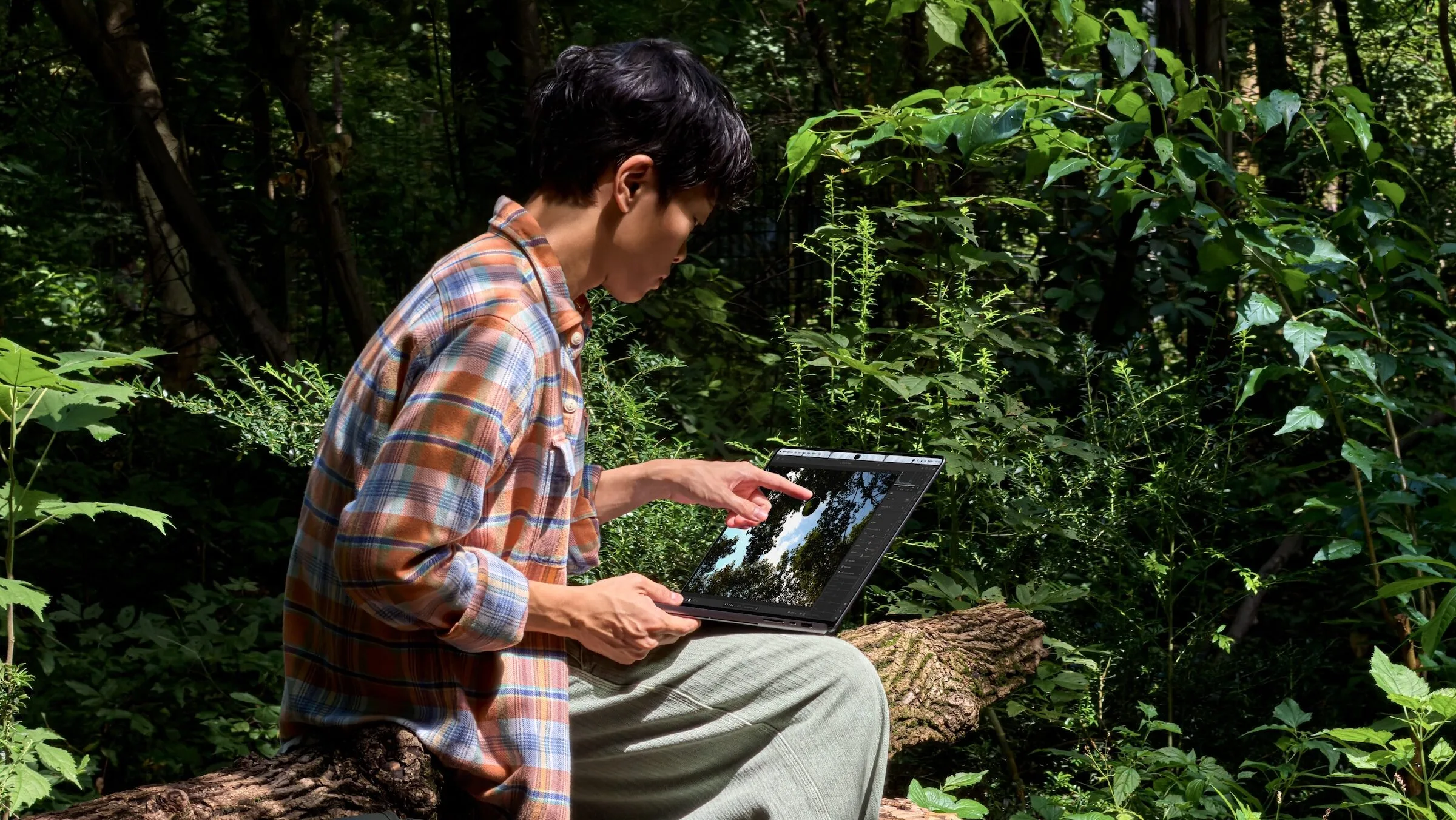 Mann bearbeitet im Wald auf einem Baumstamm ein Foto per Touch auf dem MacBook Pro mit PROLED XDR Display, neben ihm eine Kamera