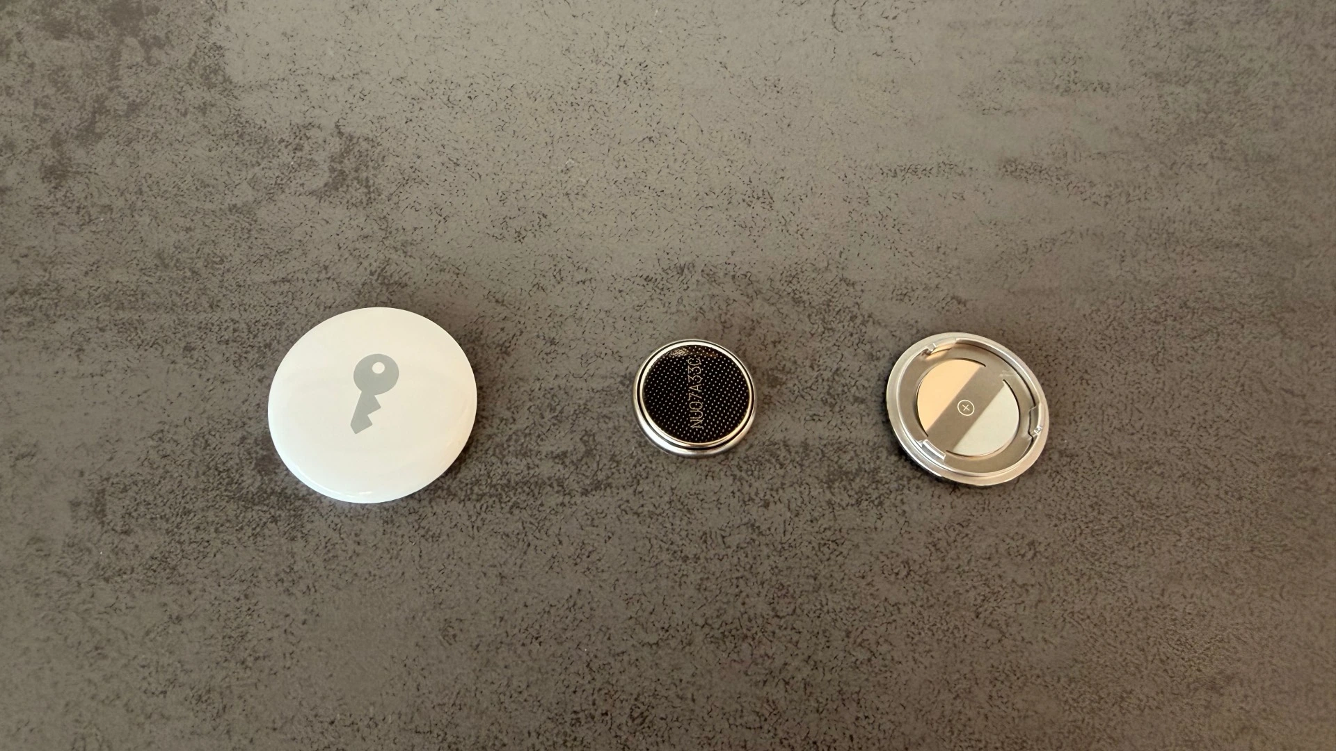 Geöffneter AirTag mit Gehäuse links, Knopfzelle in der Mitte und Abdeckung rechts, das Entnehmen der Batterie beendet AirTag-Stalking sofort
