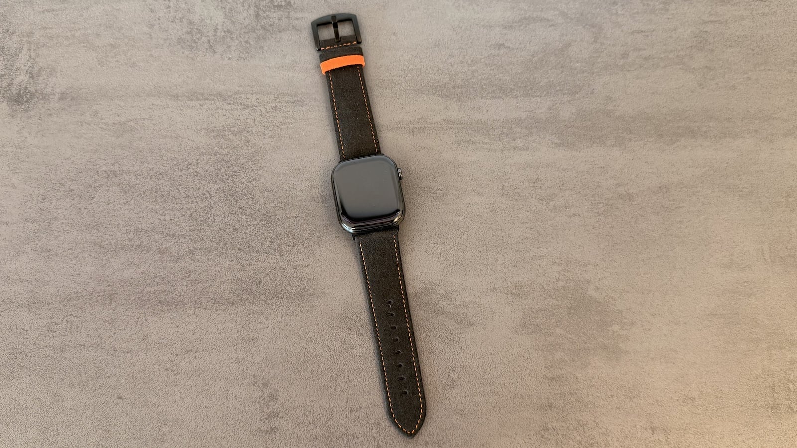 Apple Watch on the strap from the Alcantara set, laid out with the pin buckle and orange keeper loop visible