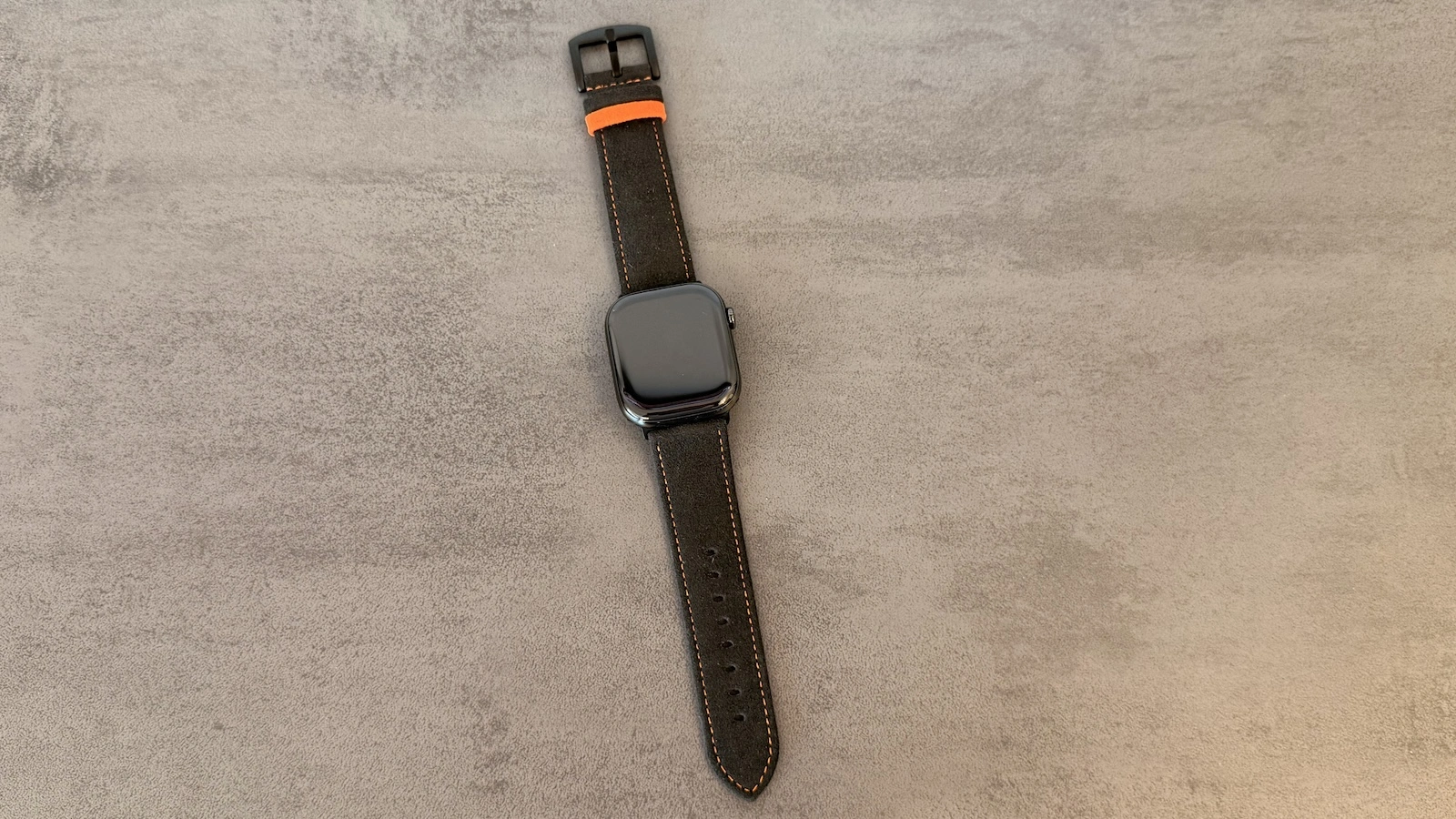 Apple Watch am Armband aus dem Alcantara Set, ausgelegt mit sichtbarer Dornschließe und orangefarbenem Halteschlaufen-Detail