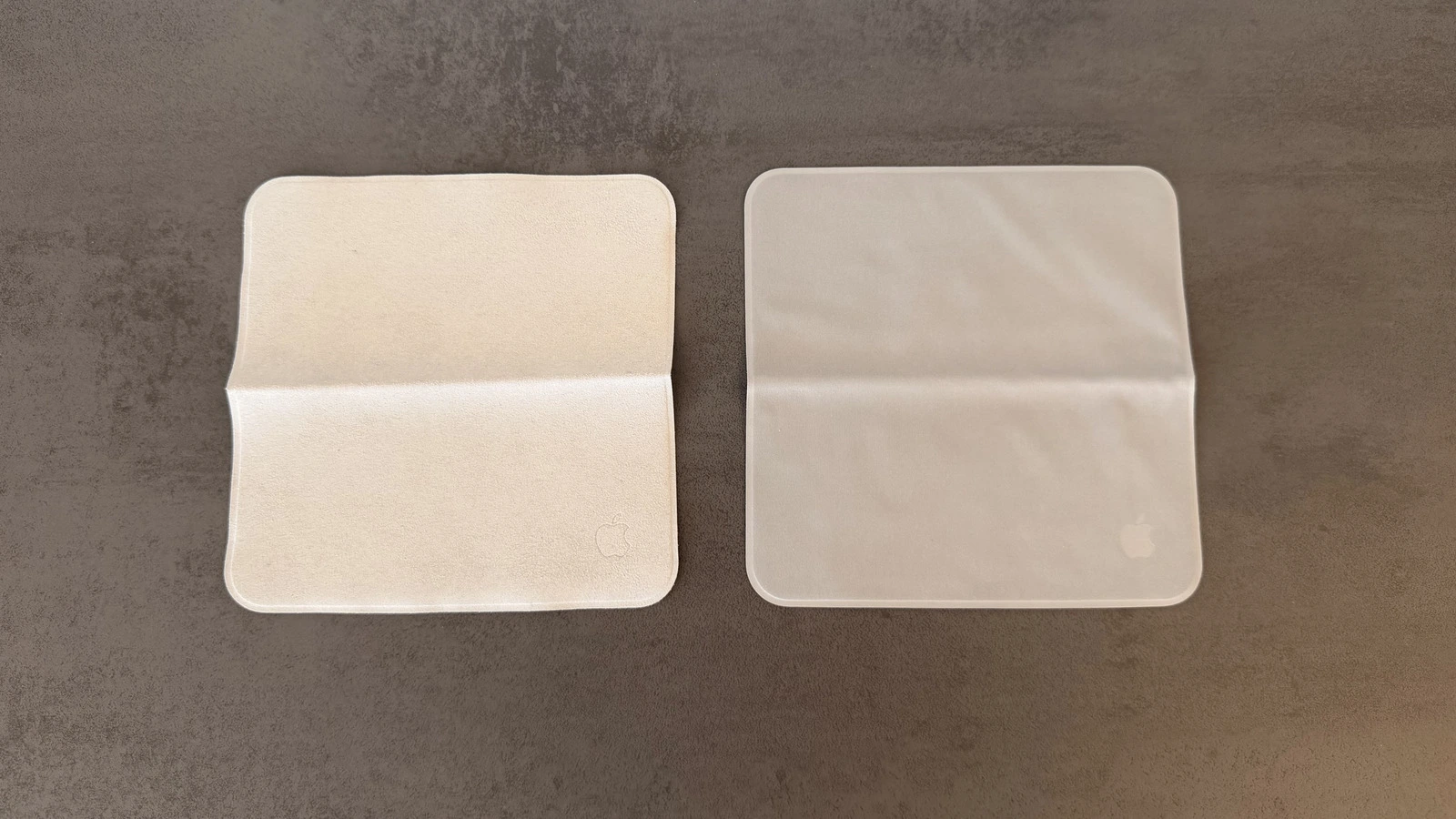 Zwei Apple Poliertücher nebeneinander auf grauem Untergrund, links die Fassung von 2021 in warmem Beige, rechts die Version von 2026 in kühlerem Grauton