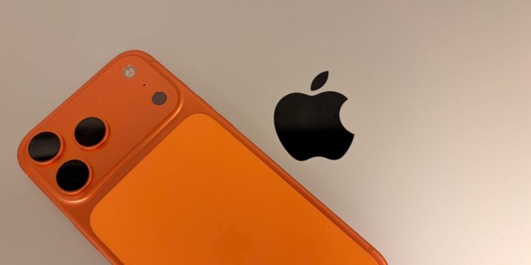 An iPhone 17 Pro Max in Cosmic Orange resting on a silver MacBook Pro next to the Apple logo, the two devices to secure when travelling with an iPhone