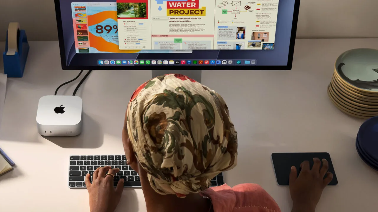 Mac mini M6 neben einem Studio Display auf einem Schreibtisch, davor eine Person an Tastatur und Trackpad