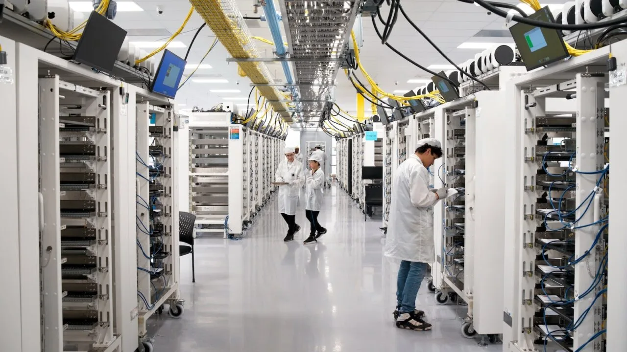 Apple KI-Server: Gasse zwischen Server-Racks in Apples Werk in Houston, Mitarbeitende in Schutzkleidung prüfen die Geräte