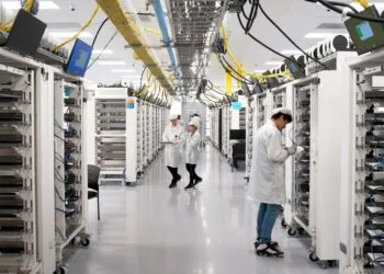 Apple KI-Server: Gasse zwischen Server-Racks in Apples Werk in Houston, Mitarbeitende in Schutzkleidung prüfen die Geräte