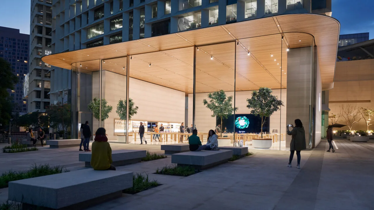 Apple Store Genius Bar: Außenansicht eines Apple Store mit Glasfront, Holzdach und Plaza mit Steinbänken am Abend