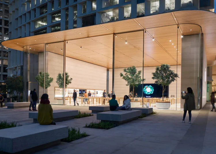 Apple Store Genius Bar: Außenansicht eines Apple Store mit Glasfront, Holzdach und Plaza mit Steinbänken am Abend