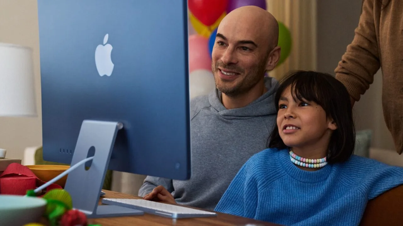 Ein Kind sitzt mit einem Erwachsenen vor einem blauen iMac, Motiv zu Apples Leitfaden für Bildschirmzeit in Familien