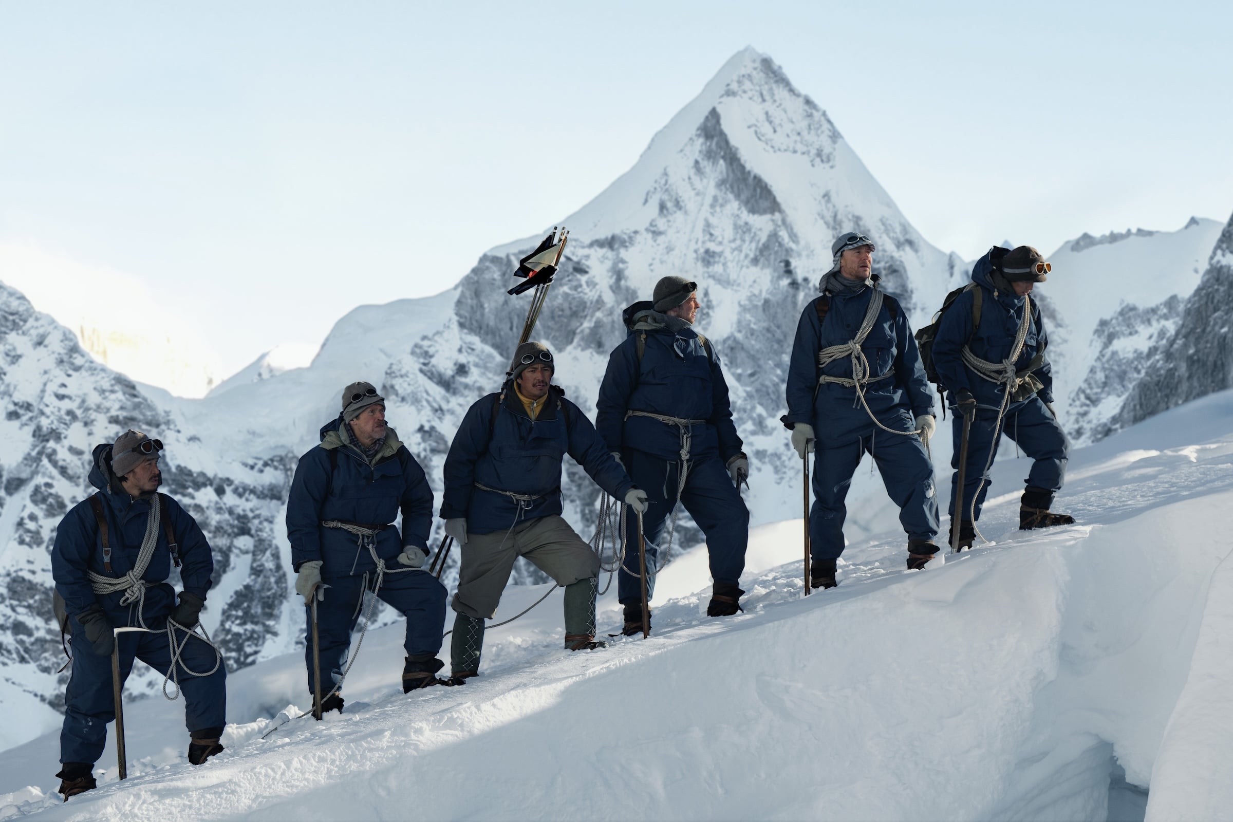 Apple TV October 2026: six roped mountaineers in blue suits stand on a snow ridge in front of a pointed peak, scene from Tenzing