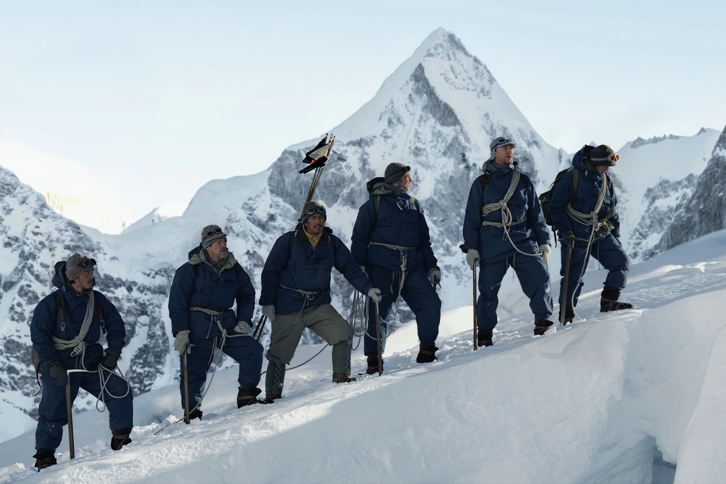 Apple TV Oktober 2026: Sechs Bergsteiger in blauen Anzügen stehen angeseilt auf einem Schneegrat vor einem spitzen Gipfel, Szene aus Tenzing
