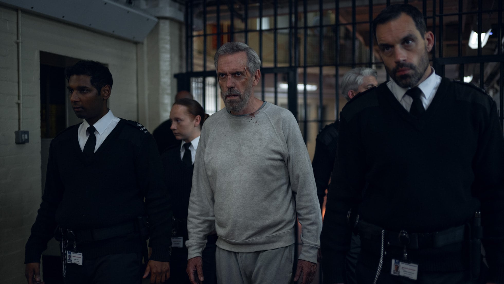Hugh Laurie as Felix Carmichael in a grey tracksuit between prison officers, a scene from The Wanted Man on Apple TV