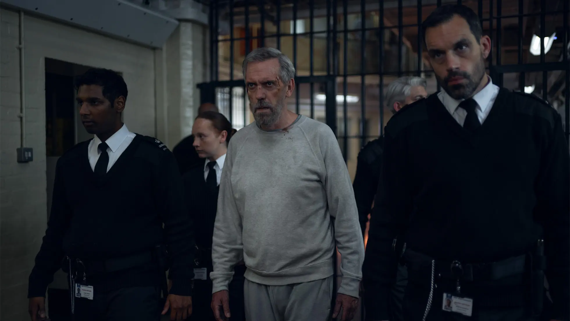 Hugh Laurie als Felix Carmichael in grauem Trainingsanzug zwischen Vollzugsbeamten im Gefängnis, Szene aus The Wanted Man bei Apple TV