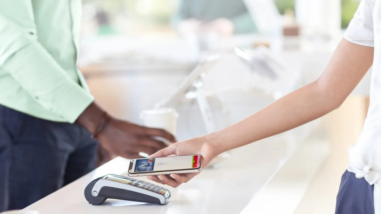 Apple Pay NFC Großbritannien: Eine Hand hält ein iPhone mit abgeschlossener Apple-Pay-Zahlung über ein Kartenterminal auf einer Ladentheke