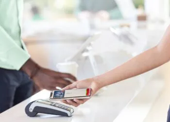 Apple Pay NFC Großbritannien: Eine Hand hält ein iPhone mit abgeschlossener Apple-Pay-Zahlung über ein Kartenterminal auf einer Ladentheke