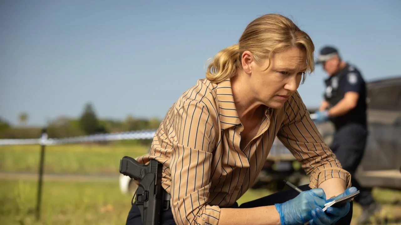 Szenenbild aus Dustfall bei Apple TV: Anna Torv als Ermittlerin Tig Pollard kniet mit Notizblock und Handschuhen an einem abgesperrten Tatort, im Hintergrund ein Polizist