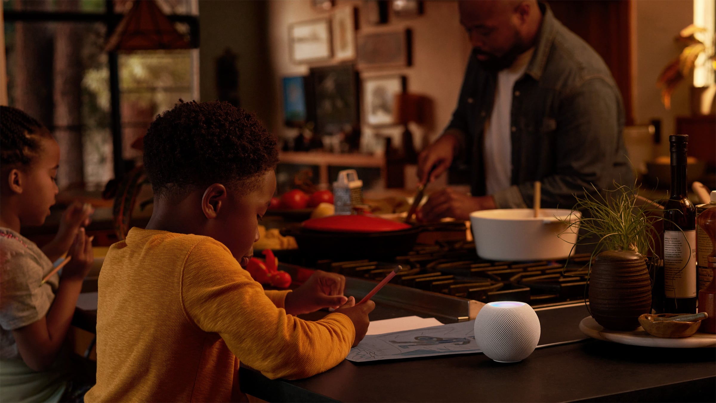 HomePod mini vs. HomePod: a white HomePod mini with an illuminated top on a kitchen counter, two children drawing beside it and a man cooking at the stove in the background