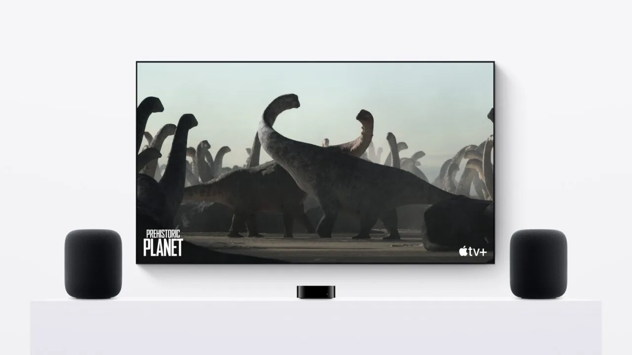 HomePod Stereopaar: Zwei HomePods links und rechts eines Fernsehers, in der Mitte ein Apple TV auf einem weißen Sideboard