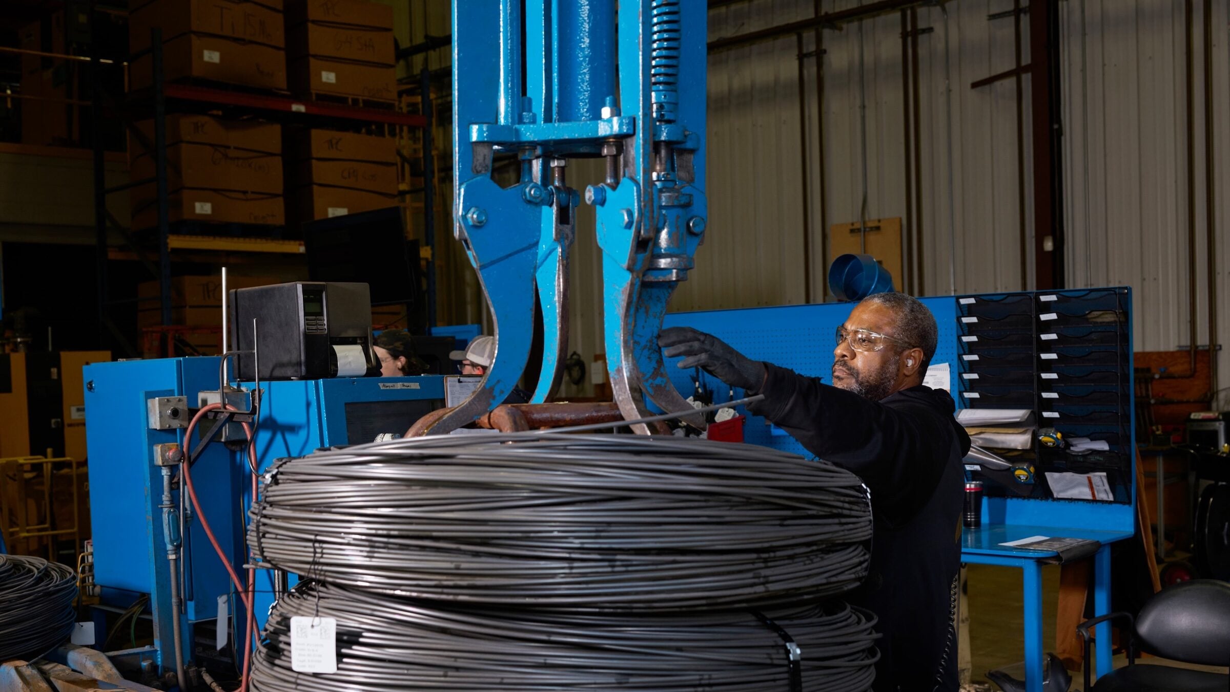 iPhone Duo titanium USA: worker wearing safety glasses positions a gripper on stacked coils of metal wire in a factory hall