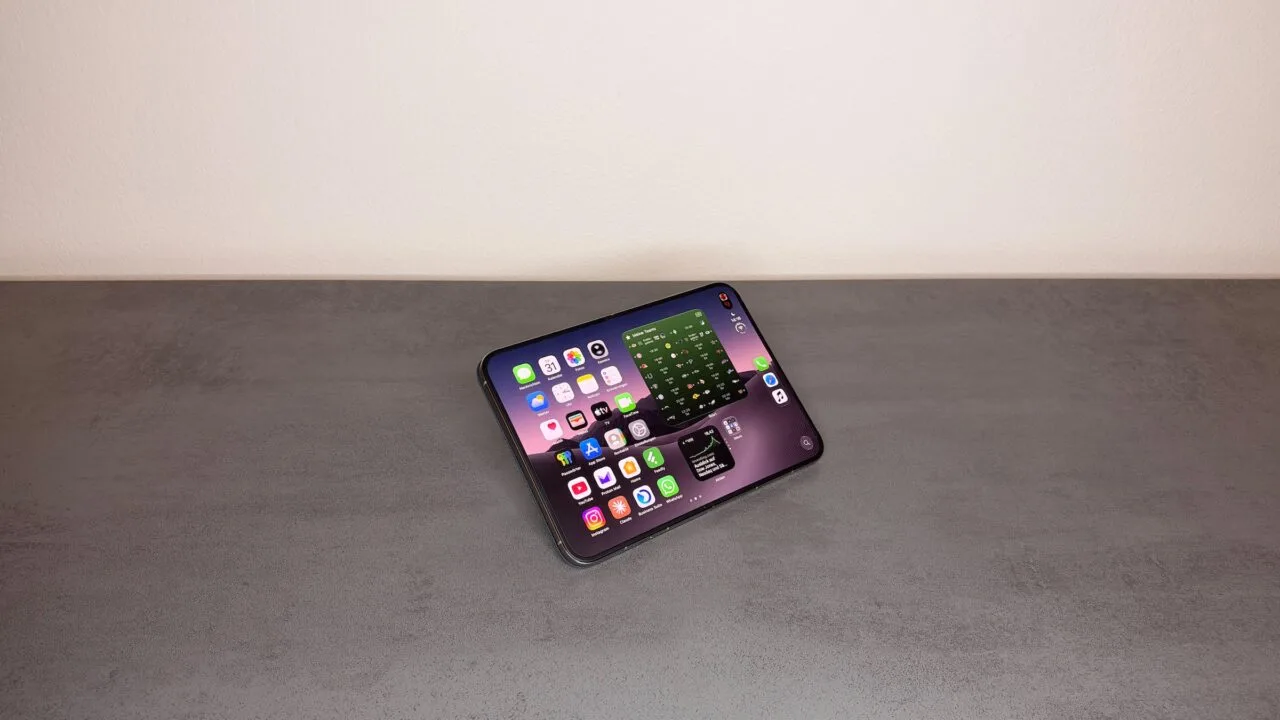 iPhone Duo Widgets auf dem Home-Bildschirm: aufgeklapptes iPhone Duo im Querformat auf einem grauen Tisch, links App-Symbole, rechts Widgets mit Spielplan und Börsenkurs