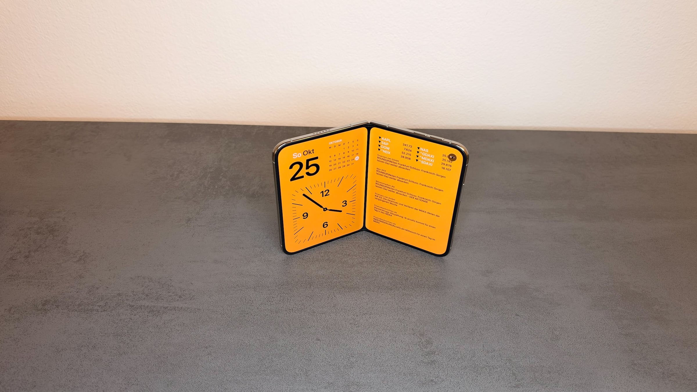 iPhone Duo StandBy in book position: the half-open iPhone Duo stands on a table, its inner display showing the date, a calendar, an analog clock and stock prices.