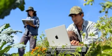 Ein Mann arbeitet zwischen Pflanzen an einem silbernen MacBook Pro M6, dahinter notiert eine Frau etwas auf einem iPad