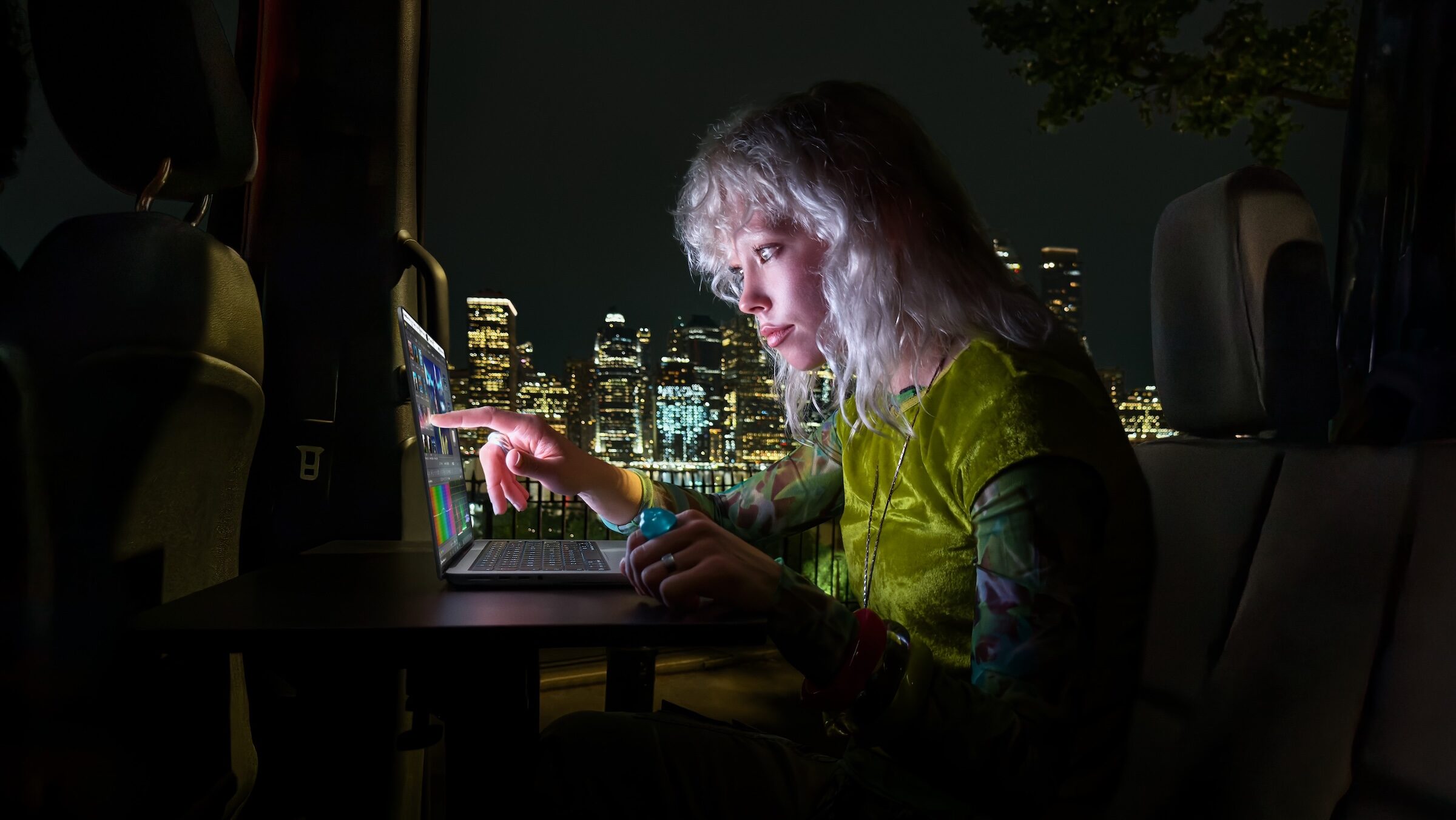 Woman tapping the display of a MacBook Pro with PROLED XDR Display with her finger inside a van at night, a lit-up skyline behind her