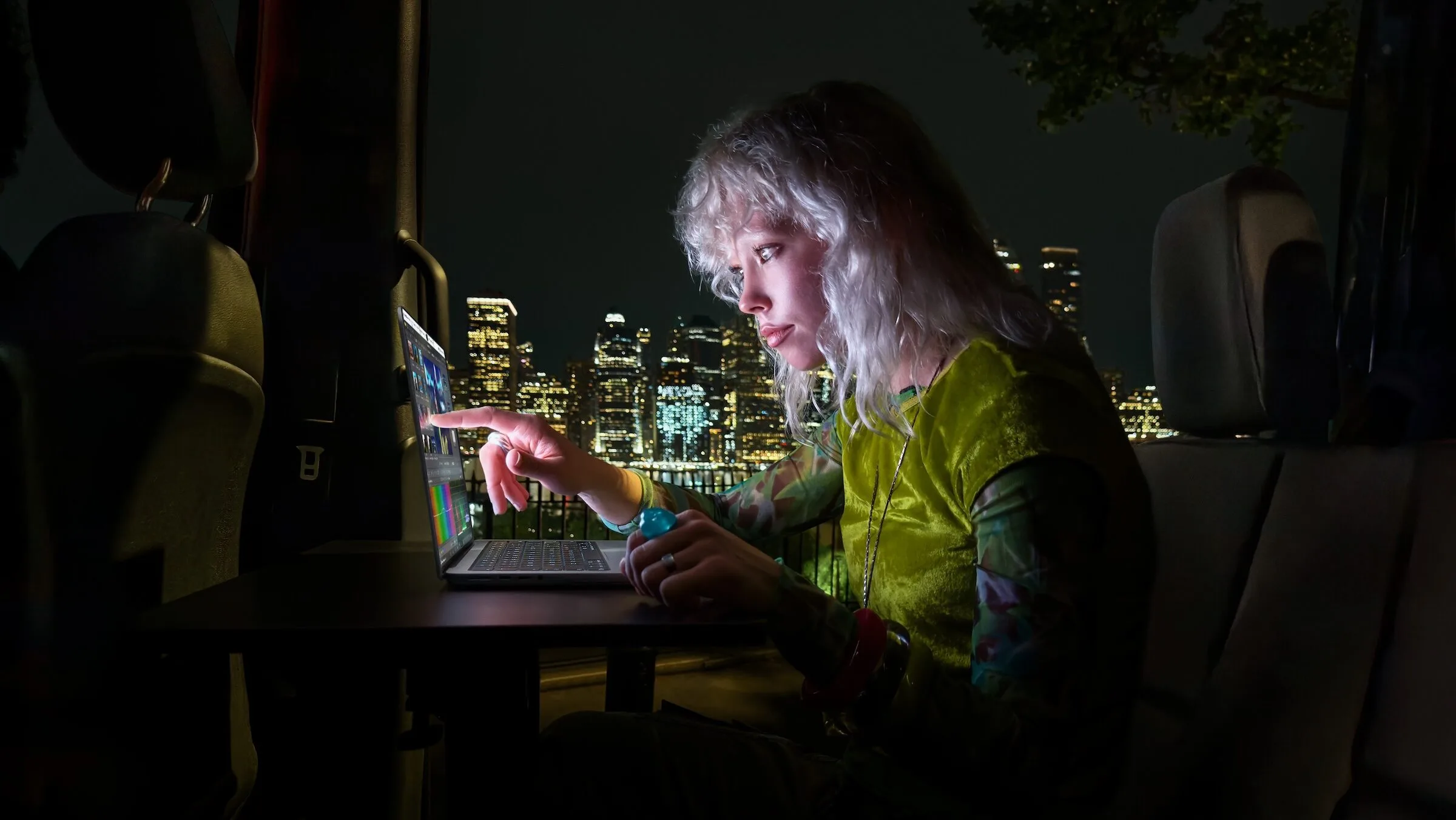 Frau tippt nachts in einem Kleinbus mit dem Finger auf das Display eines MacBook Pro mit PROLED XDR Display, dahinter eine beleuchtete Skyline
