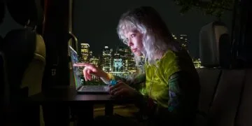 MacBook Pro Touch: Frau tippt nachts in einem Kleinbus mit dem Finger auf das Display eines MacBook Pro mit PROLED XDR Display, dahinter eine beleuchtete Skyline
