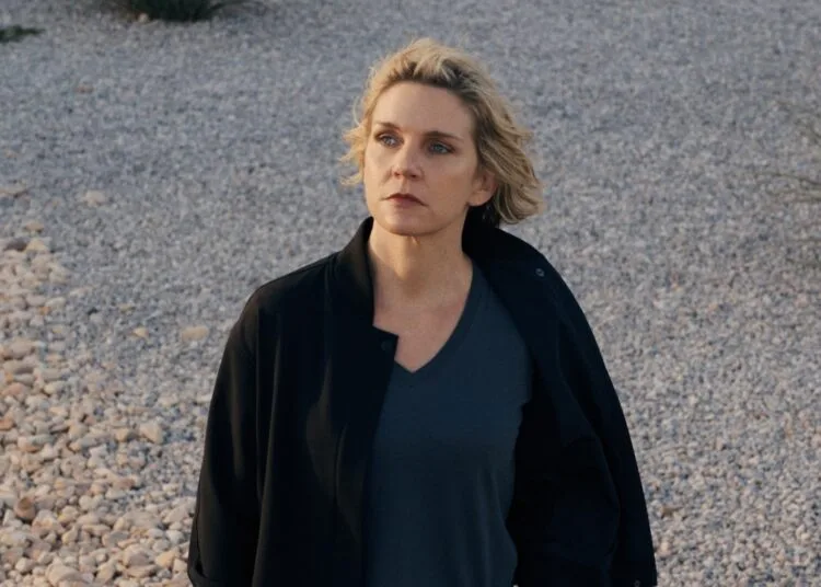 Pluribus Staffel 2: Rhea Seehorn als Carol Sturka steht in schwarzer Jacke auf einer Kiesfläche mit Wüstenpflanzen, Szene aus der ersten Staffel