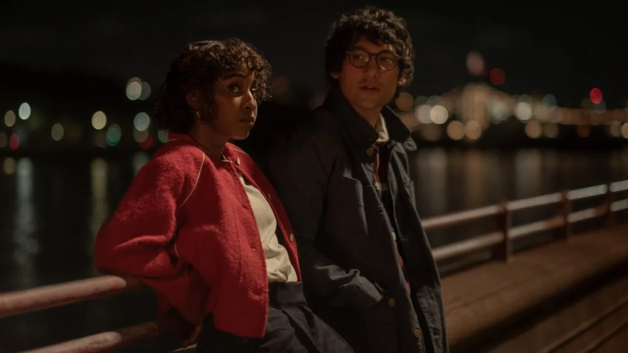 Szenenbild aus Prodigies: Ayo Edebiri in roter Jacke und Will Sharpe mit Brille lehnen nachts an einem Geländer am Wasser, im Hintergrund Lichter einer Stadt