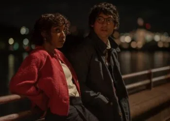 Szenenbild aus Prodigies: Ayo Edebiri in roter Jacke und Will Sharpe mit Brille lehnen nachts an einem Geländer am Wasser, im Hintergrund Lichter einer Stadt