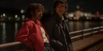 Szenenbild aus Prodigies: Ayo Edebiri in roter Jacke und Will Sharpe mit Brille lehnen nachts an einem Geländer am Wasser, im Hintergrund Lichter einer Stadt