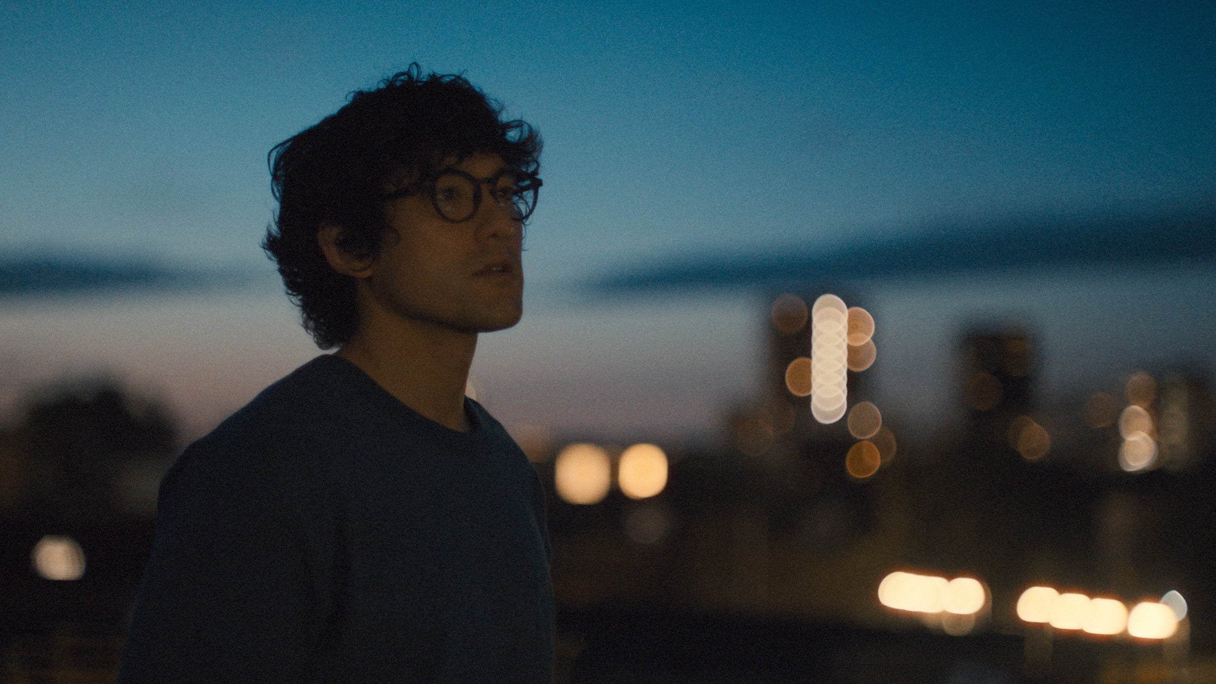 Still from Prodigies: Will Sharpe in glasses at dusk, blurred city lights in the background