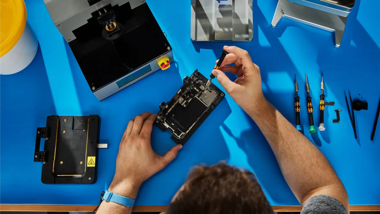 Self Service Repair iPhone 18 Pro: Hände reparieren ein geöffnetes iPhone in einer Halterung auf einer blauen Werkbank, daneben Apples Reparaturgeräte und Schraubendreher