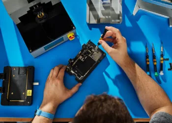 Self Service Repair iPhone 18 Pro: Hände reparieren ein geöffnetes iPhone in einer Halterung auf einer blauen Werkbank, daneben Apples Reparaturgeräte und Schraubendreher