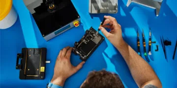 Self Service Repair iPhone 18 Pro: Hände reparieren ein geöffnetes iPhone in einer Halterung auf einer blauen Werkbank, daneben Apples Reparaturgeräte und Schraubendreher