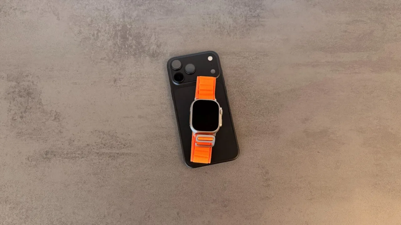 Taptic Engine Lizenzgebühren: schwarzes iPhone mit Kamerafeld, darauf eine Apple Watch Ultra mit orangefarbenem Alpine Loop auf grauem Steintisch