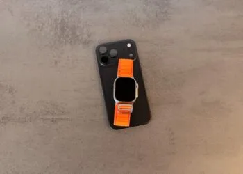 Taptic Engine Lizenzgebühren: schwarzes iPhone mit Kamerafeld, darauf eine Apple Watch Ultra mit orangefarbenem Alpine Loop auf grauem Steintisch