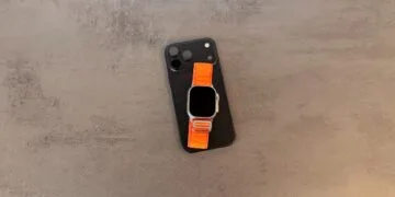 Taptic Engine Lizenzgebühren: schwarzes iPhone mit Kamerafeld, darauf eine Apple Watch Ultra mit orangefarbenem Alpine Loop auf grauem Steintisch