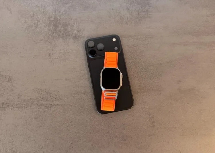 Taptic Engine Lizenzgebühren: schwarzes iPhone mit Kamerafeld, darauf eine Apple Watch Ultra mit orangefarbenem Alpine Loop auf grauem Steintisch