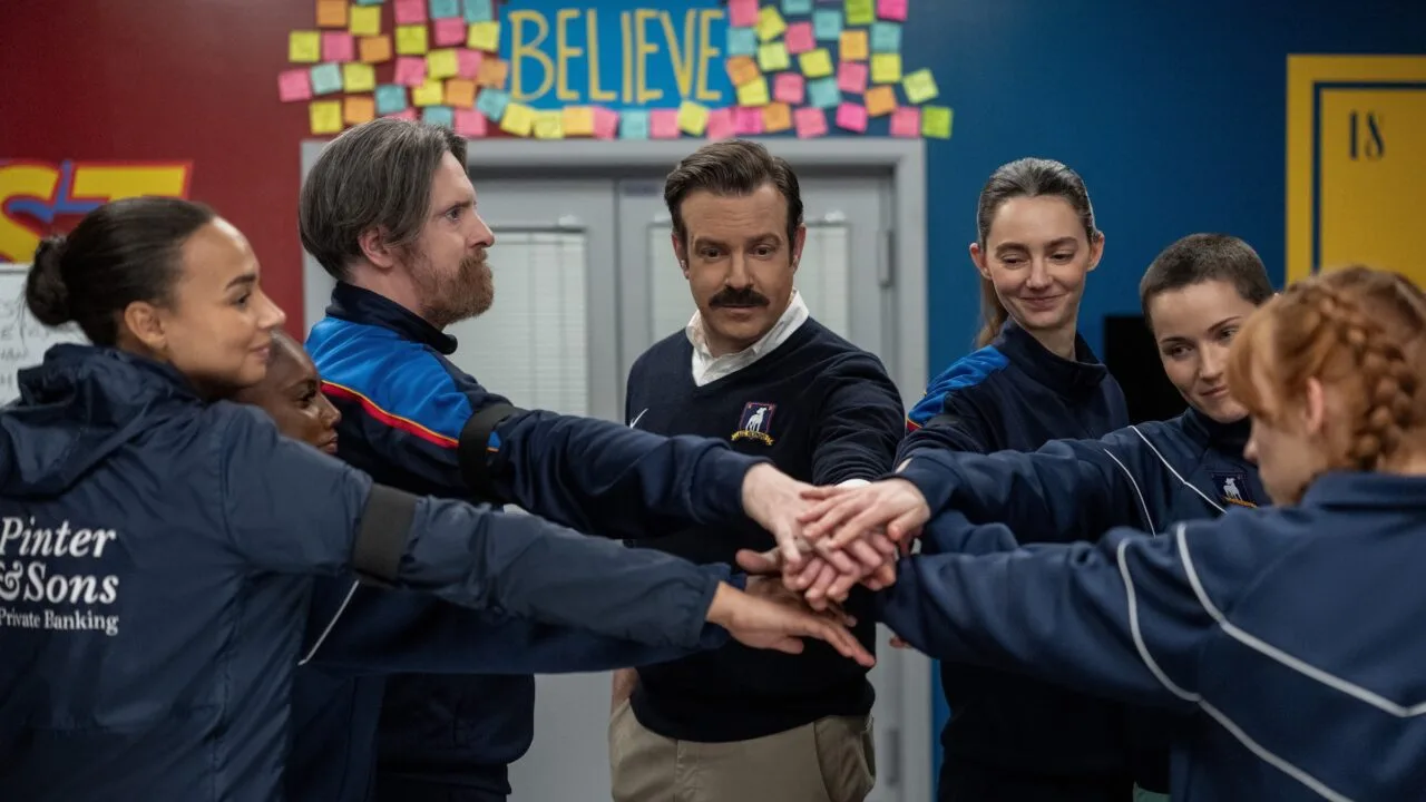 Ted Lasso Staffel 5: Ted Lasso legt mit dem Frauenteam von AFC Richmond die Hände zusammen, darüber das Believe-Schild