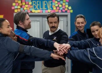 Ted Lasso Staffel 5: Ted Lasso legt mit dem Frauenteam von AFC Richmond die Hände zusammen, darüber das Believe-Schild